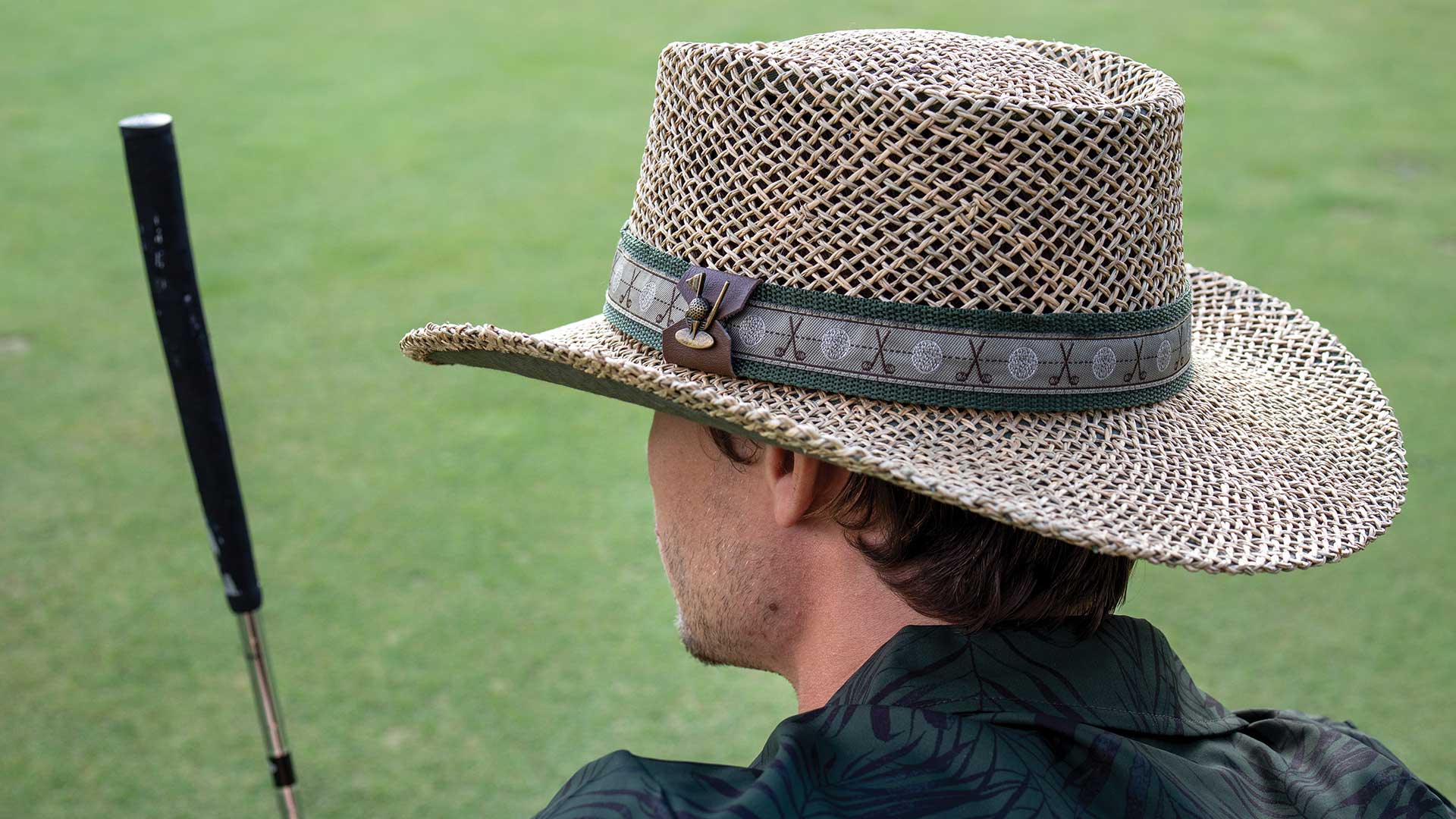 Man wearing straw hat with golf club and golf ball pattern while carrying a putter.