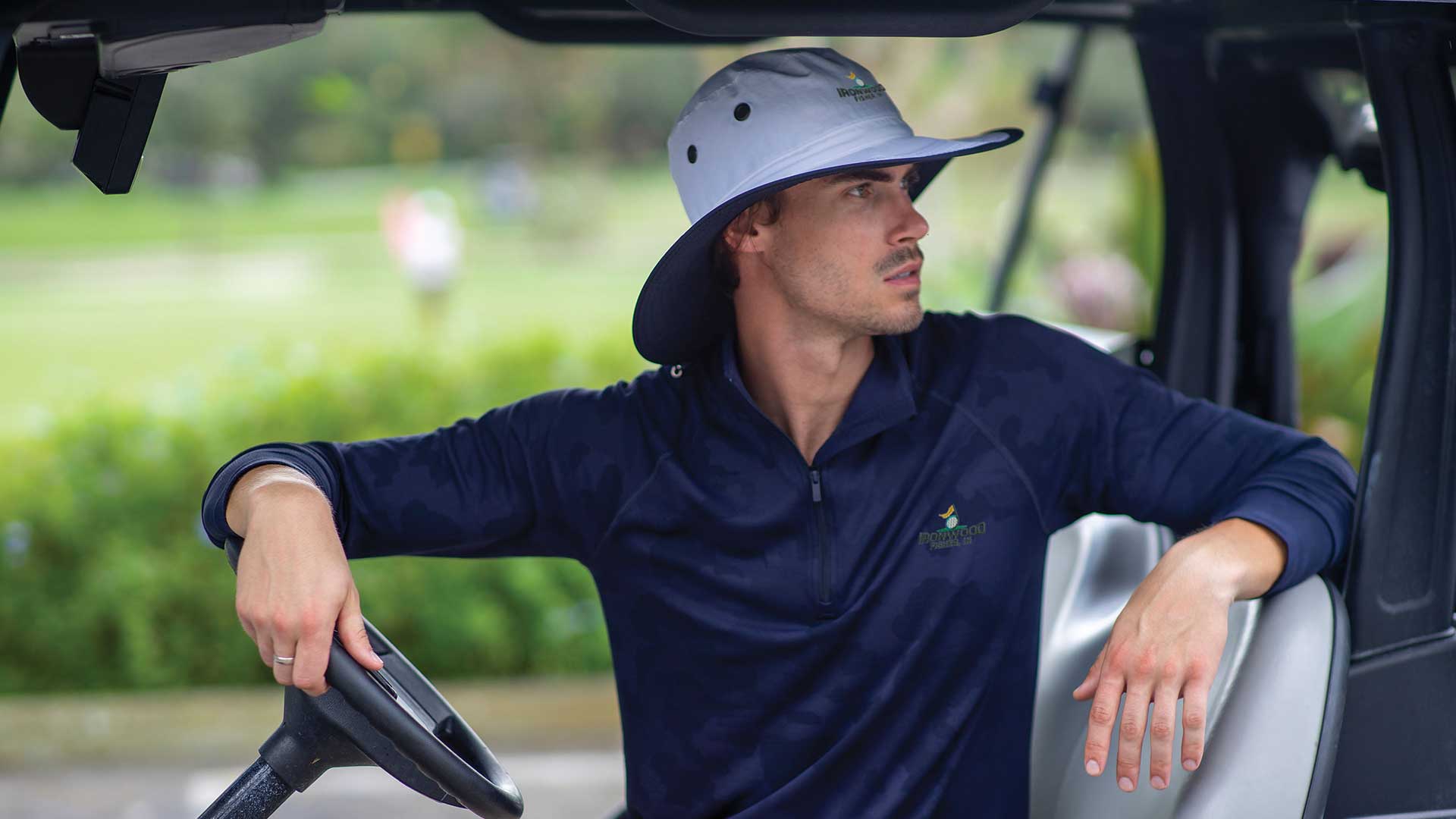 Man wearing a hat in a golf cart with long blue sleeve embroidered shirt.